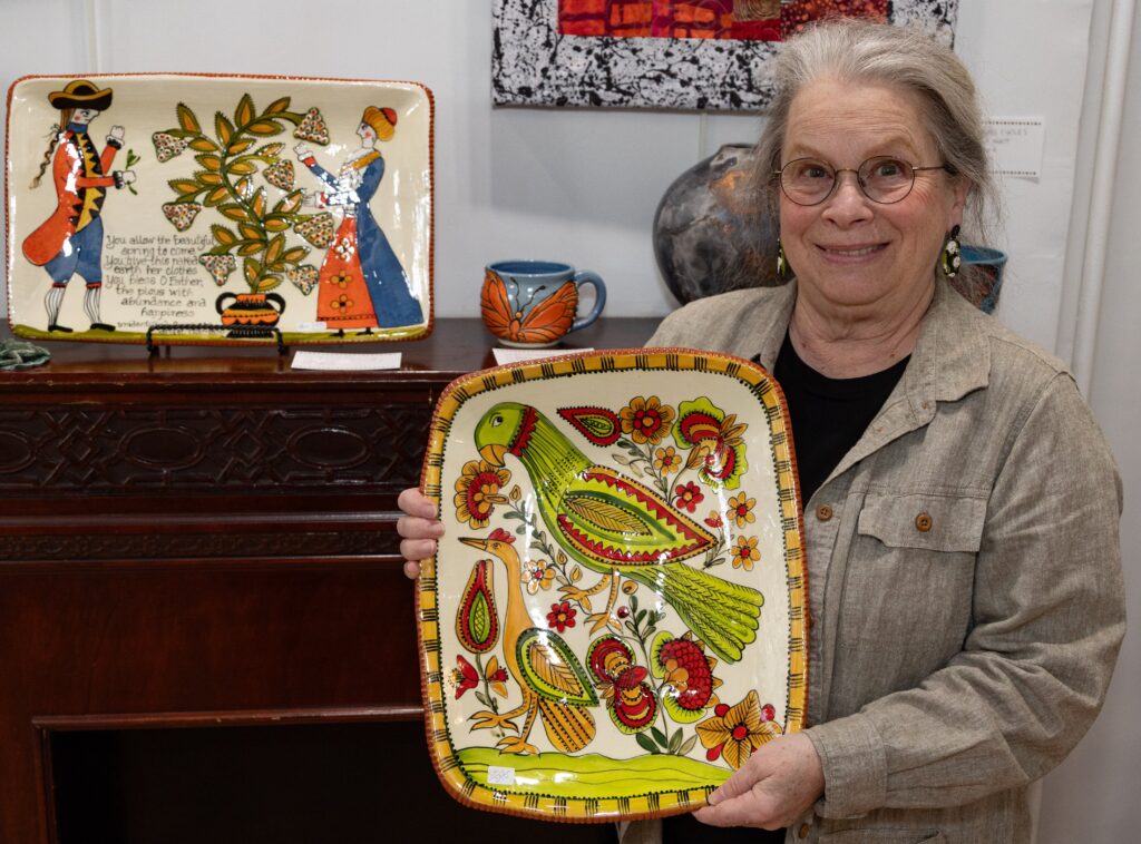 Artist Selinda Kennedy posing with redware platter featuring the design of a folk art parrot and flowers in green and red colors