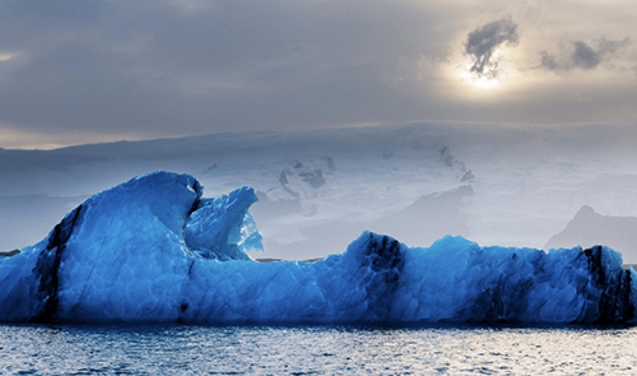 Photograph by Owen Floody. A brilliant blue iceberg floats in choppy waters.  Inland, steep ridges rise to the late-afternoon sunset sky.